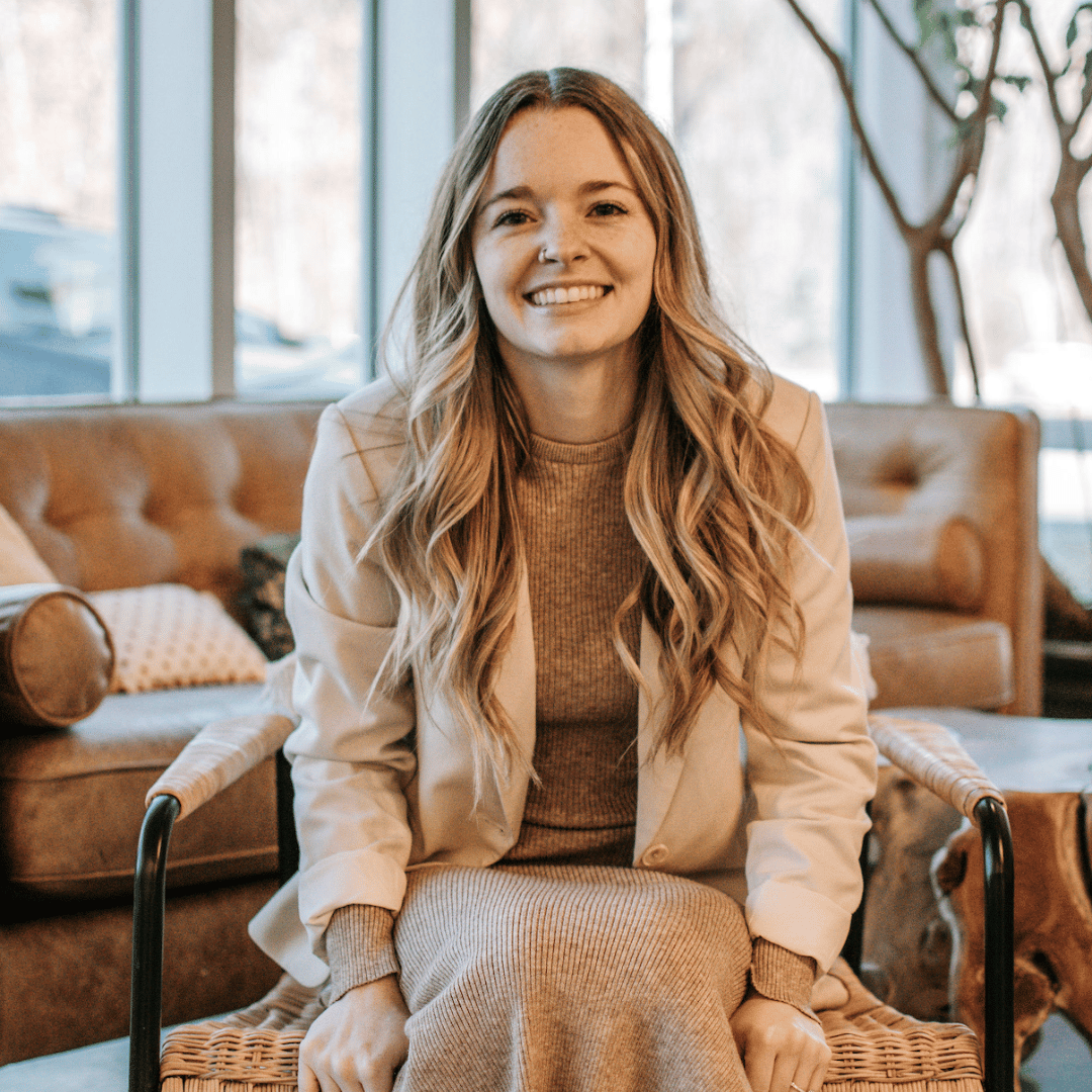 Chelsea sits in a chair smiling, with a brown leather sofa and windows in the background.