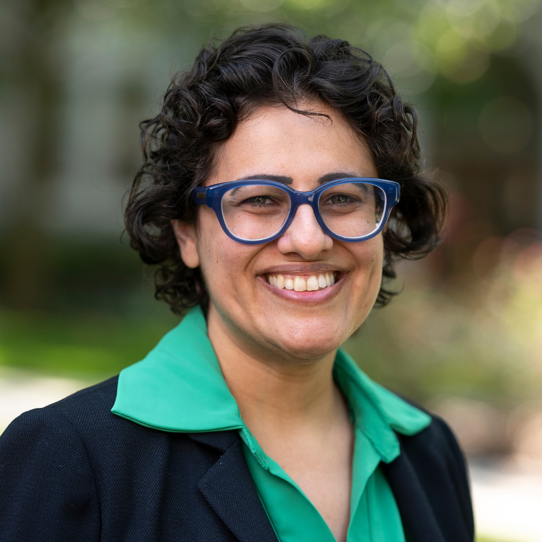 Marina smiles in a green shirt and black blazer, wearing blue framed eyeglasses with Loyola's evergreen campus in the back.