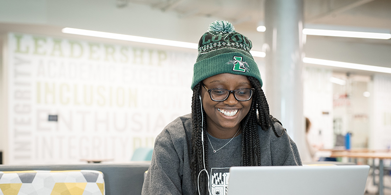 Student smiling and looking at a computer while wearing a Loyola hat
