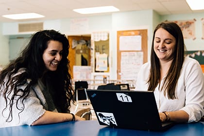 Two smiling teachers sitting in a classroom and looking at a laptop