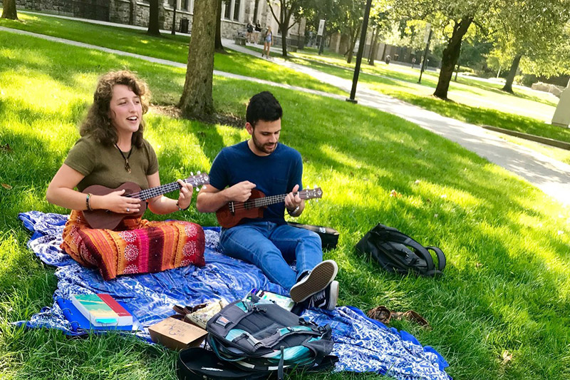Two students playing ukeleles on blankets on grass