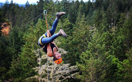 A student smiling and hanging upside down on a zipline over a heavily wooded area