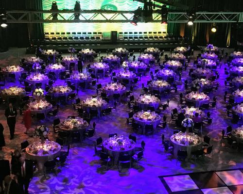 Wide angle shot of a large event hall set with dozens of round tables and purple event lighting