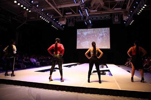 4 female models side-by-side on the runway in the same hands-on-hips pose with backs to the camera.