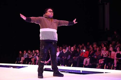 A male model standing alone at the end of the runway in a color-block outfit with arms stretched out to the sides.